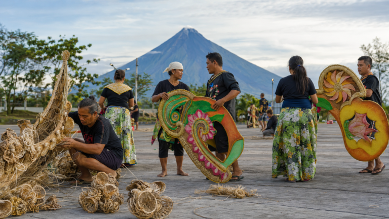 Adult Bicolano festival makers preparing abaca-based props with Mayon Volcano in the distance